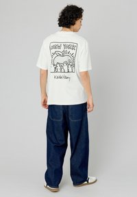 T-shirt blanc avec un design graphique noir représentant une pomme et des mains, avec le texte "NEW YORK" et "Keith Haring". Coupe ample, manches courtes.