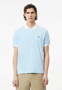 Polo tričko v svetlom modrom odtieni s bielymi pruhmi, krátkymi rukávmi a bielym golierom. Na ľavej hrudi sa nachádza zelené logo Lacoste.