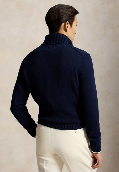 Homme de dos portant un pull à col roulé côtelé bleu marine et un pantalon couleur crème sur un fond neutre.