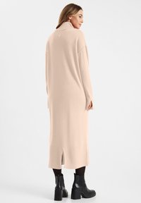 Lange, losvallende beige gebreide jurk met een hoge kraag, zijsplitten en geribbelde accenten aan de zoom. Geraadpleegd met zwarte platform enkellaarsjes.