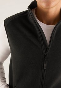 Sort fleecevest med høj krave og lynlås hele vejen foran, med glat tekstur og diskrete syningsdetaljer. Ensfarvet hvid skjorte under.