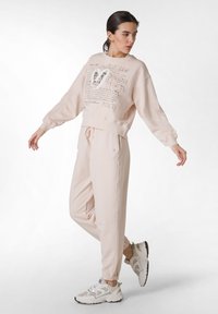 Ljusrosa sweatshirt med grafisk text, tillsammans med matchande joggare. Har en avslappnad passform, elastiska ärmslut och vita sneakers med detaljer.