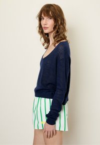 Tamsiai mėlynas megztinis su V formos iškirpte, laisvo kirpimo ir ilgomis rankovėmis, kartu su baltais šortais, turinčiais žalias vertikalias juosteles.