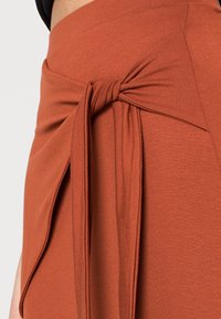 Jupe portefeuille couleur rouille en tissu doux, avec un détail noué et un drapé sur le côté pour plus de texture et de style.