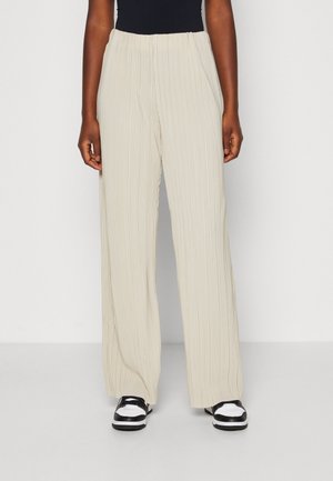 Person wearing beige wide-leg pleated pants and black-and-white sneakers, standing against a white background.