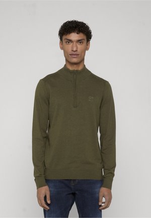 Homme aux cheveux bouclés portant un pull à demi-fermeture éclair vert olive et un jean bleu, debout devant un fond neutre.