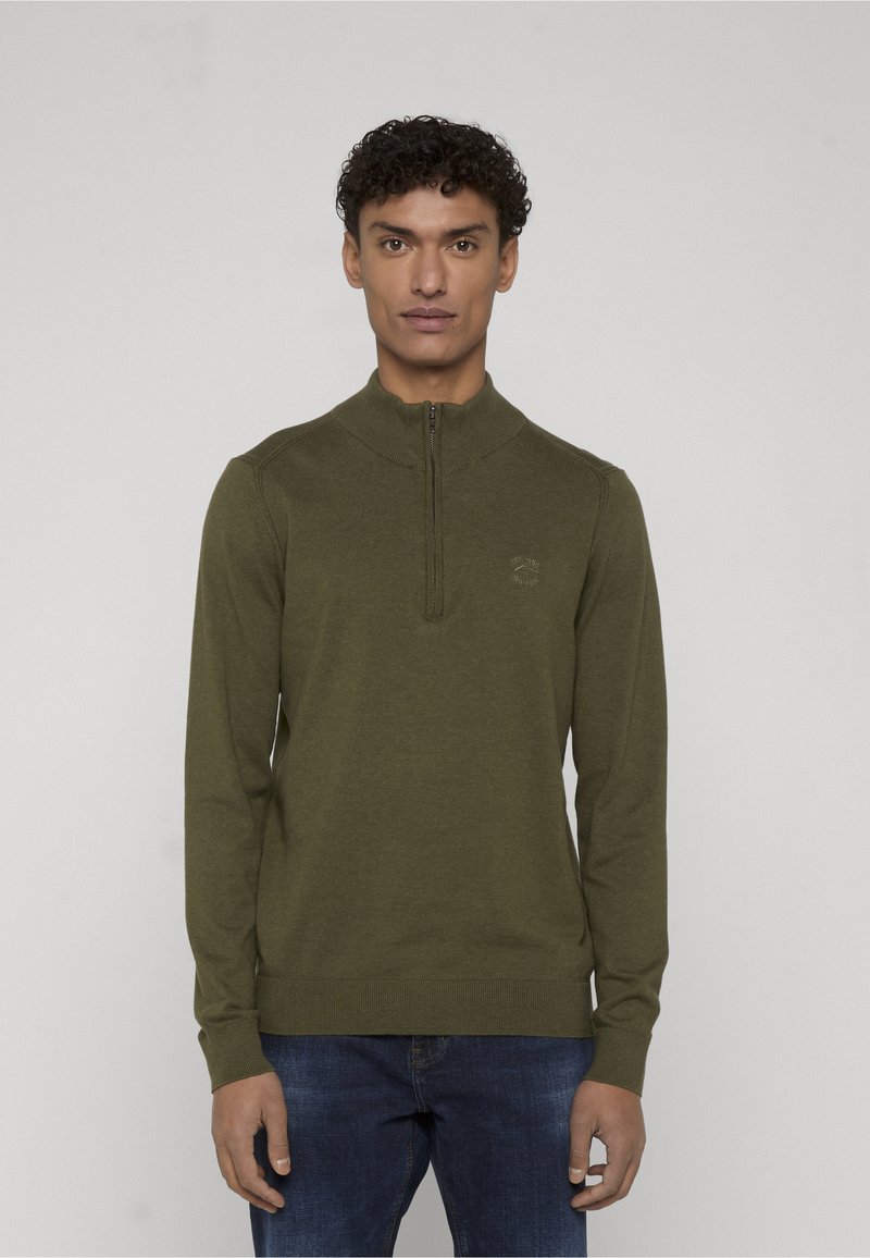 Homme aux cheveux bouclés portant un pull à demi-fermeture éclair vert olive et un jean bleu, debout devant un fond neutre.