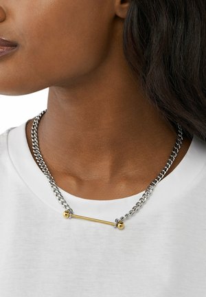 Silverfärgad kedjehalsband med ett horisontellt guldbars och två runda gulddetaljer, bärs över en vit skjorta. Slät textur, modern design.