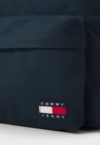 Mörkblå tygväska med en framträdande rektangulär textilapplikation som har orden "TOMMY JEANS" och en röd, vit och blå design.