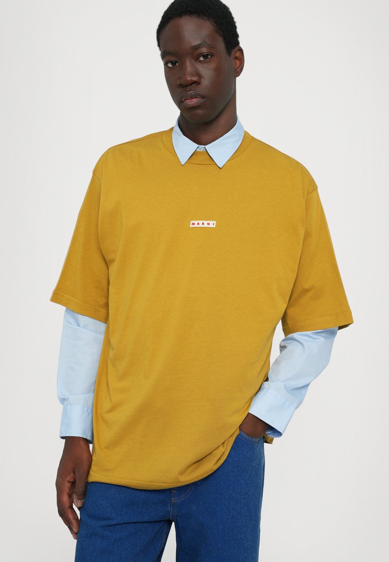 Mustard yellow oversized t-shirt with a crew neck, featuring a small red and white logo patch. Light blue dress shirt visible underneath.