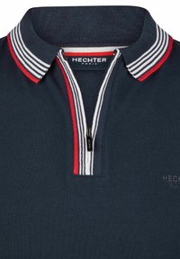 Polo shirt navy avec col zippé, doté d'accents rayés rouges et blancs. Le tissu est texturé, avec un branding discret sur la poitrine.