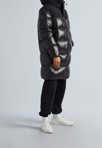 Svart puffjacka med glänsande yta, knälång, med huva, dragkedjestängning och framfickor. Bärs med svarta byxor och vita sneakers.