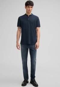 Polo shirt bleu marine à manches courtes avec un col henley, associé à un jean en denim noir et des chaussures noires à lacets, sur un fond gris uni.