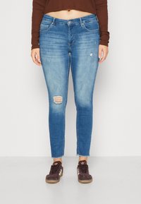 Světle modré skinny džíny se zvlněnými detaily, včetně trhliny na pravém koleni. Mají střední pas a frené spodní lem, spárované s hnědými teniskami.