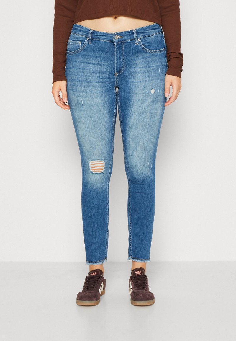 Světle modré skinny džíny se zvlněnými detaily, včetně trhliny na pravém koleni. Mají střední pas a frené spodní lem, spárované s hnědými teniskami.