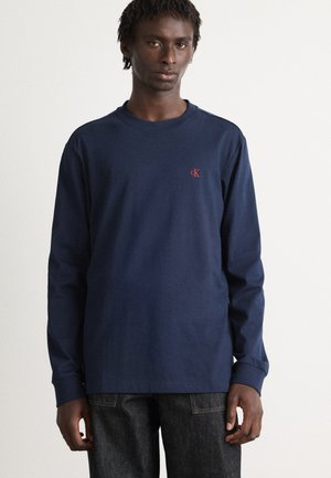 Jonge man in een marineblauw longsleeve shirt met een klein rood "cK" logo op de linkerborst, staande tegen een effen lichte achtergrond.