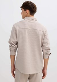 Camisa beige clara con tejido texturizado, mangas largas, puños abotonados y detalle de canesú trasero, con dobladillo curvo y ajuste casual.