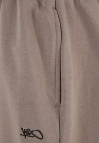 Lysbrune sweatpants lavet af blødt stof, med sidelommer og et sort broderet logo på venstre ben. Elastisk talje inkluderet.
