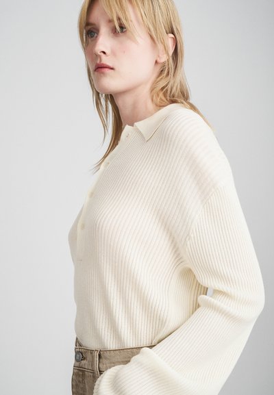 Cream ribbed knit sweater with a collared neckline and button placket. Features relaxed fit and long sleeves, paired with khaki pants.