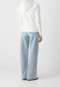 Felpa bianca con cappuccio, in un tessuto morbido, abbinata a jeans a gamba larga di colore blu chiaro, dotati di tasche posteriori e una finitura sbiadita.