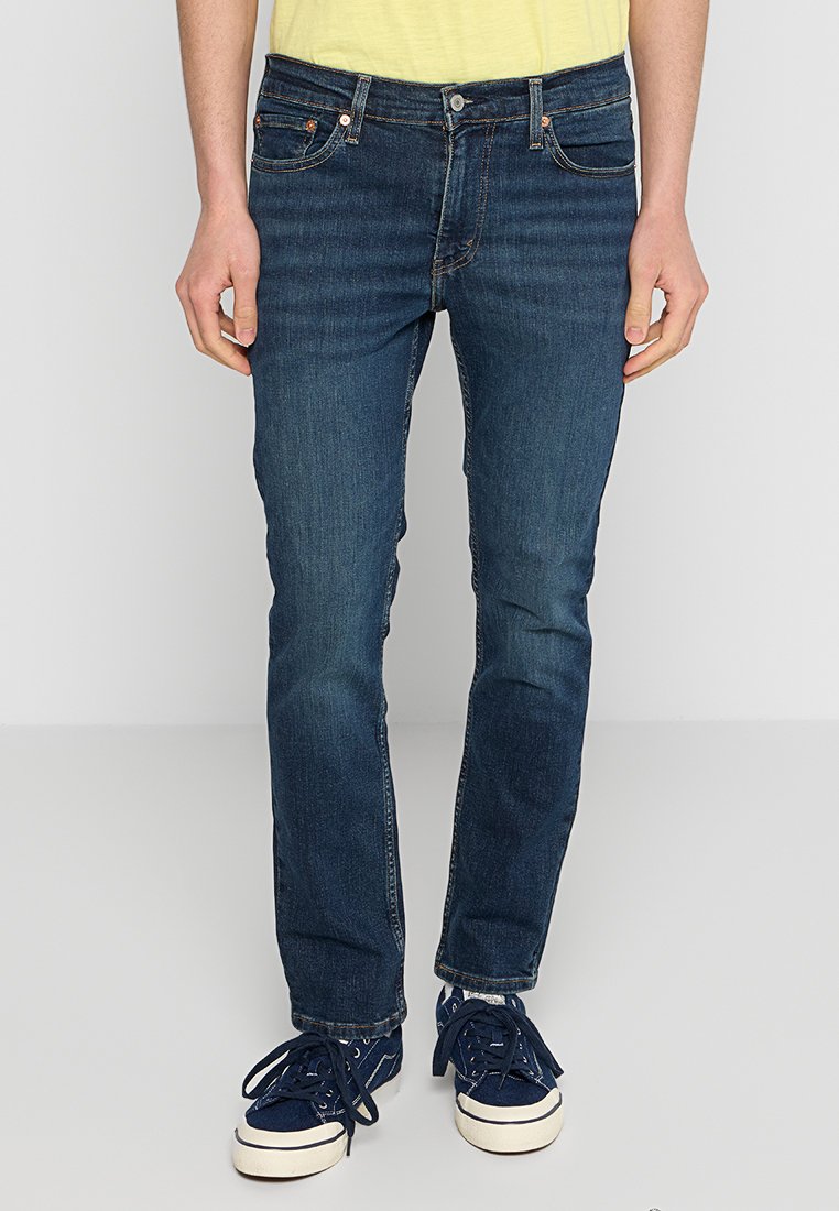 Dunkelblaue Jeans aus Denim im Slim-Fit, mit einem Fünf-Taschen-Design, kontrastierenden Nähten und einem Knopfverschluss. Getragen mit navyblauen Sneakers.