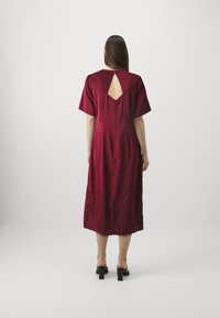 Burgundijos atlaso suknelė su trumpomis rankovėmis, iki kelių ilgio, ir rakto skylute nugaros dalyje. Lygi tekstūra ir elegantiškas krintančio audinio efektas. Juodos blokinės kulno šlepetės.
