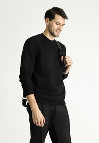 Felpa nera in materiale morbido con scollatura rotonda e polsini a coste. Indossata con pantaloni neri, caratterizzati da un dettaglio con spallaccio.