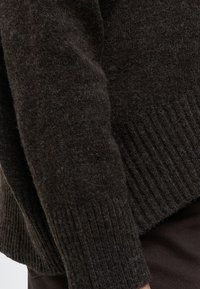 Gros plan d'une manche de pull en laine brun foncé avec des poignets côtelés portée sur un pantalon foncé.
