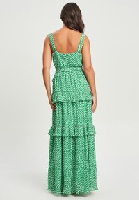 Maxi obleka iz zelene tkanine z belim cvetličnim vzorcem, ki ima nabrané plasti in široke naramnice, zasnovana z oprijetim korzetom in razširjenim krilom.