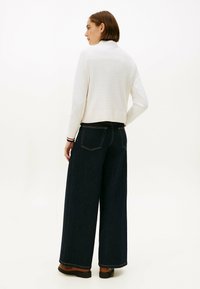 Pull en maille texturée blanche associé à un jean large bleu foncé. L'ensemble est complété par des bottines marron et des détails de couture visibles.