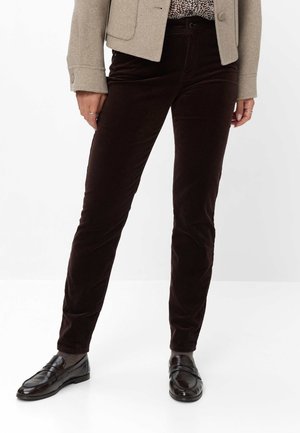 Vrouw draagt donkerbruine fluwelen broek, lichtbeige jas, blouse met patroon, grijze sokken en zwarte leren loafers, staand tegen een witte achtergrond.