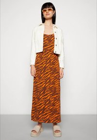 Mulher vestida com um vestido maxi em padrão de zebra laranja e castanho, jaqueta cropped branca, sandálias plataforma bege e óculos de sol âmbar, a posar contra uma parede branca.