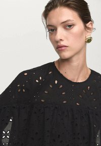 Haut noir avec des découpes en œillets complexes, un col rond et un design à volants superposés le long de l'ourlet. Texture subtile sur l'ensemble.