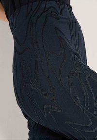 Close-up van donkere broek met glanzende, golvende borduursels en een achterzak zichtbaar tegen een lichte achtergrond.