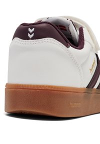 Hvide sneakers med en maroon hæl og striber, tekstureret gummisål og velcro-rem. Har branding på siden og en glat overflade.