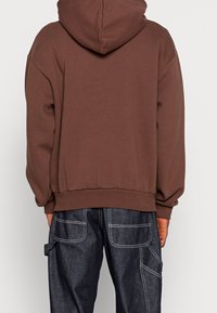 Brun luvtröja med avslappnad passform, ribbade muddar och nederkant, i kombination med mörka denimbyxor med kontrasterande vit söm.