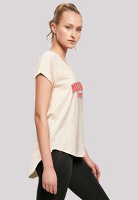 Beige short-sleeve T-shirt with a rounded neckline featuring bold red text. The shirt has a relaxed fit and extends slightly at the back.