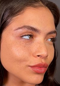 Närbild av ett ansikte med slät hy, fräknar, hasselbruna ögon, mörkbrunt hår och matt mauve läppstift. Subtila högdagrar på kinderna.