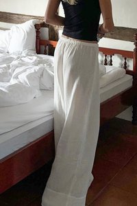 Valged, laia lõikega, tekstuuriga püksid, mis on valmistatud kergest kangast, koos sobiva musta ülaosaga. Taustal on voodi, millel on kortsus valge voodipesu.