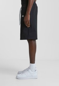 Pantalones cortos de algodón negros con cintura elástica y cordón de ajuste gris claro; presentan un parche de logo en un lado y se combinan con zapatillas blancas.