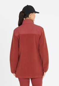 Rote Fleecejacke mit hohem Kragen und strukturiertem Stoff. Verfügt über einen kontrastierenden oberen Abschnitt und eine lässige Passform, kombiniert mit einer schwarzen Mütze.