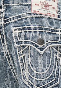 Vaqueros de mezclilla azul desgastados con costuras blancas, diseño en los bolsillos traseros y una etiqueta con un personaje de dibujos animados sosteniendo una guitarra.