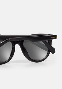 Occhiali da sole neri con finitura lucida, lenti rotonde e design elegante. Presentano il marchio dorato "Polaroid" sulla tempia.