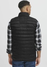 Gilet di piumino nero con sezioni a righe orizzontali, colletto alto e texture liscia, indossato sopra una camicia a maniche lunghe a scacchi.
