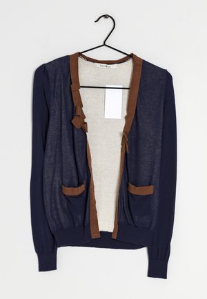 Cardigan blu navy con bordatura marrone, due tasche frontali, tessuto leggero, maniche lunghe e una texture sottile. Appeso a un gancio nero.