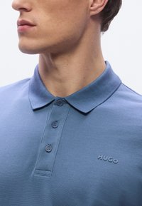 Īsām piedurknēm zilas krāsas polo krekls no teksturēta auduma, ar apkakli, trīs pogu aizdari un uzšūtu logotipu uz krūtīm.