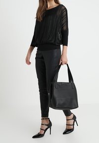 Top nero plissettato con maniche lunghe trasparenti, abbinato a pantaloni neri aderenti e tacchi neri con cinturini. Include una borsa tote nera testurizzata.