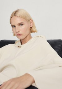 Jasnobeżowy, rozpinany sweatshirt o luźnym kroju, miękkiej teksturze i szerokich rękawach, noszony przez osobę siedzącą na czarnej skórzanej kanapie.