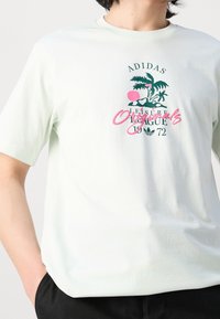 Světlezelené bavlněné tričko s grafickým designem s palmami a růžovým akcentem. Text zní "ADIDAS Leisure Originals 1972."