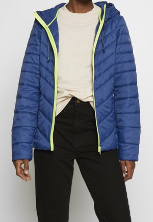 Personne portant une veste matelassée bleue avec fermeture éclair et bordure jaune néon sur un pull beige et un pantalon noir, debout devant un fond uni.